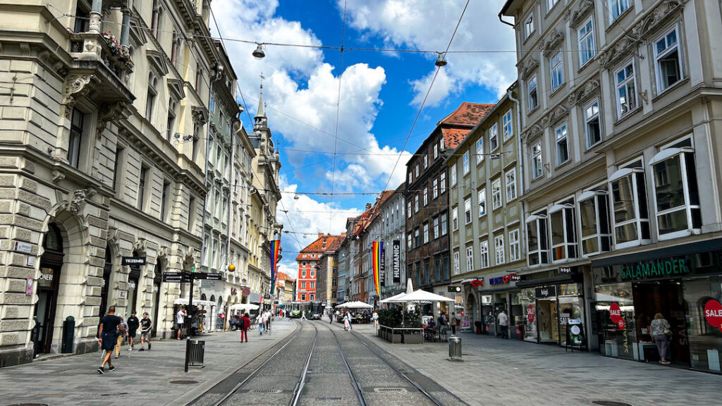 Veduta prospettica della Herrengasse a Graz, Austria, con i binari del tram che corrono al centro di una via pedonale fiancheggiata da eleganti edifici storici e negozi. Numerose persone passeggiano lungo i marciapiedi sotto un cielo azzurro punteggiato da nuvole bianche, mentre bandiere arcobaleno decorano alcune facciate.