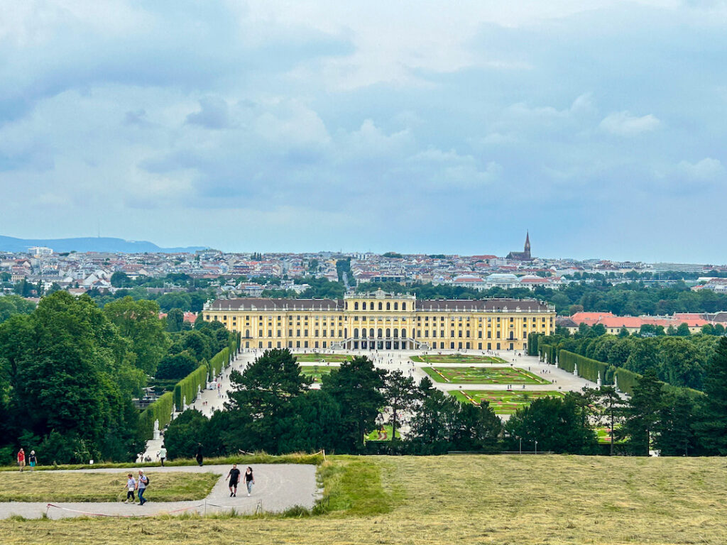 Veduta panoramica dall'alto della Reggia di Schönbrunn a Vienna, Austria. In primo piano, un prato in pendenza con visitatori che camminano su un sentiero. Al centro, l'imponente palazzo barocco giallo "Maria Teresa" con i suoi vasti giardini simmetrici e aiuole curate. Sullo sfondo, la città di Vienna si estende fino all'orizzonte sotto un cielo nuvoloso.