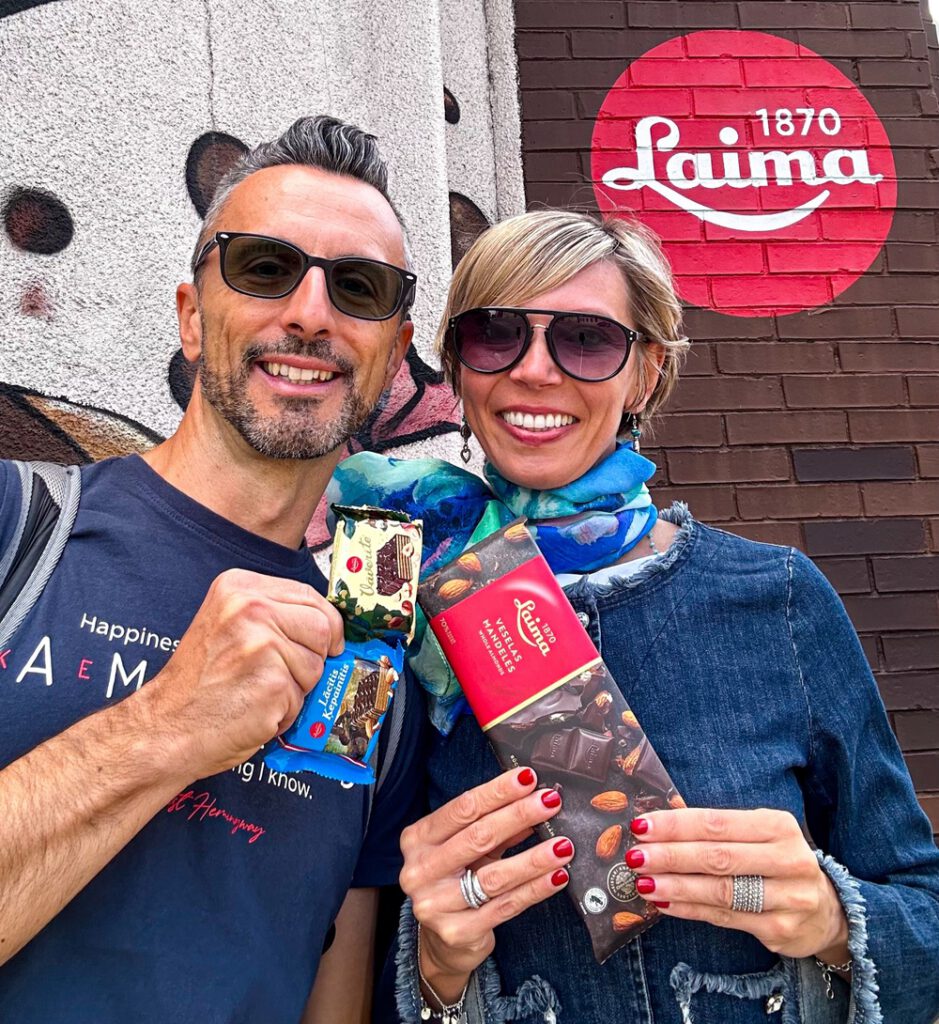 Coppia di fidanzati sorridono indossando occhiali da sole davanti al logo circolare rosso "Laima 1870" su una parete di mattoni al Museo del Cioccolato di Riga, Lettonia. Entrambi mostrano alla fotocamera diverse confezioni di cioccolato Laima, tra cui una tavoletta con mandorle e piccoli snack incartati.