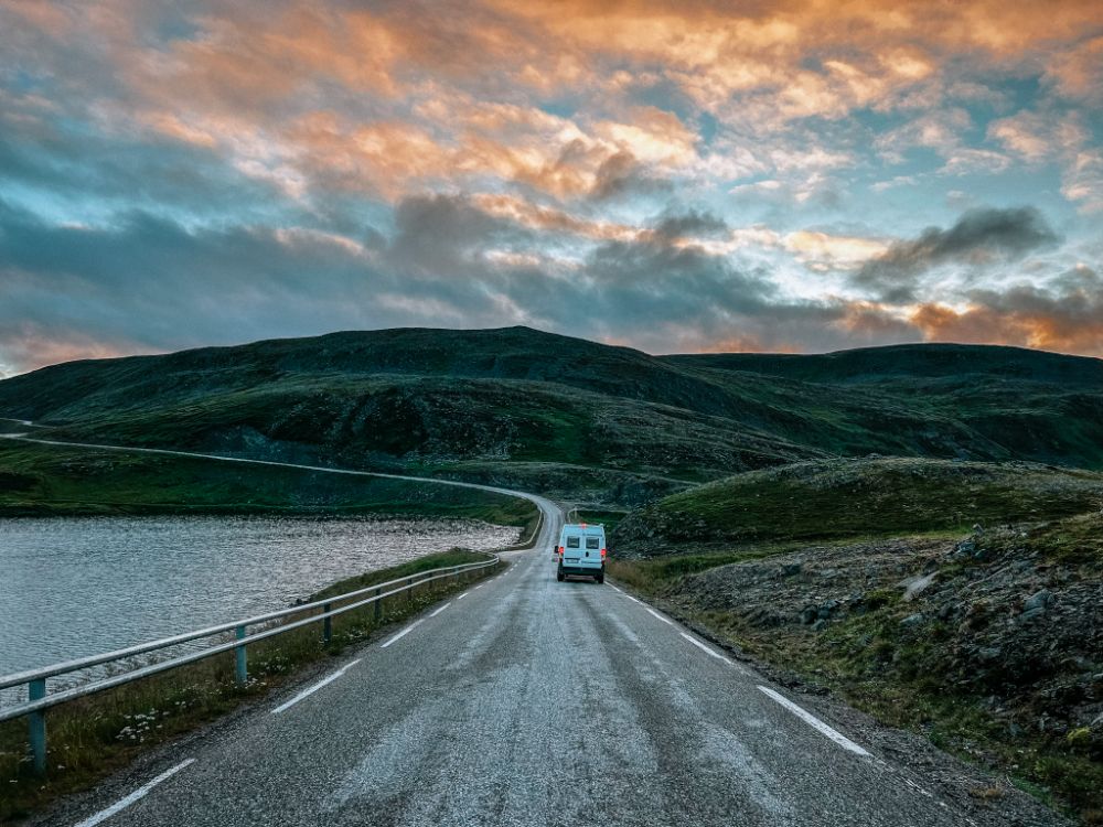 Veduta di un van bianco che percorre una strada asfaltata e sinuosa in un paesaggio montuoso e spoglio vicino a Capo Nord, Norvegia. Sulla sinistra si scorge uno specchio d'acqua, mentre il cielo è velato da nuvole illuminate dai colori caldi del tramonto estivo.