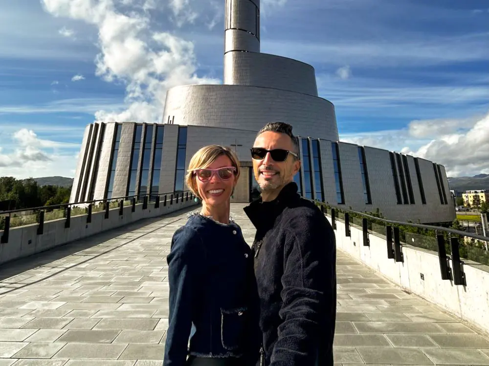 Un uomo e una donna sorridono per un selfie davanti alla Cattedrale dell'Aurora Boreale di Alta, Norvegia, un edificio moderno con una facciata metallica a spirale sotto un cielo azzurro con nuvole bianche.