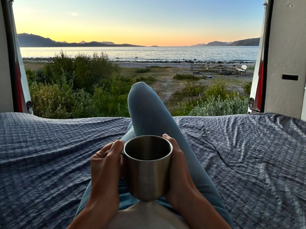Inquadratura in soggettiva dall'interno di camper/van parcheggiato sulla spiaggia di Steinsvika, Norvegia. Si vedono le mani che stringono una tazza metallica e le gambe allungate su un letto, con una vista aperta sul mare calmo, le montagne e il cielo al tramonto.