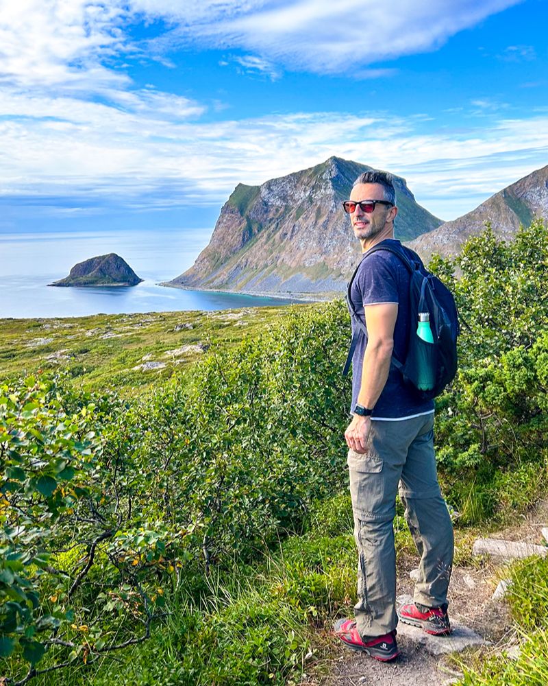Trekker sorridente con zaino su un sentiero erboso, sullo sfondo isole e mare blu alle Isole Lofoten.